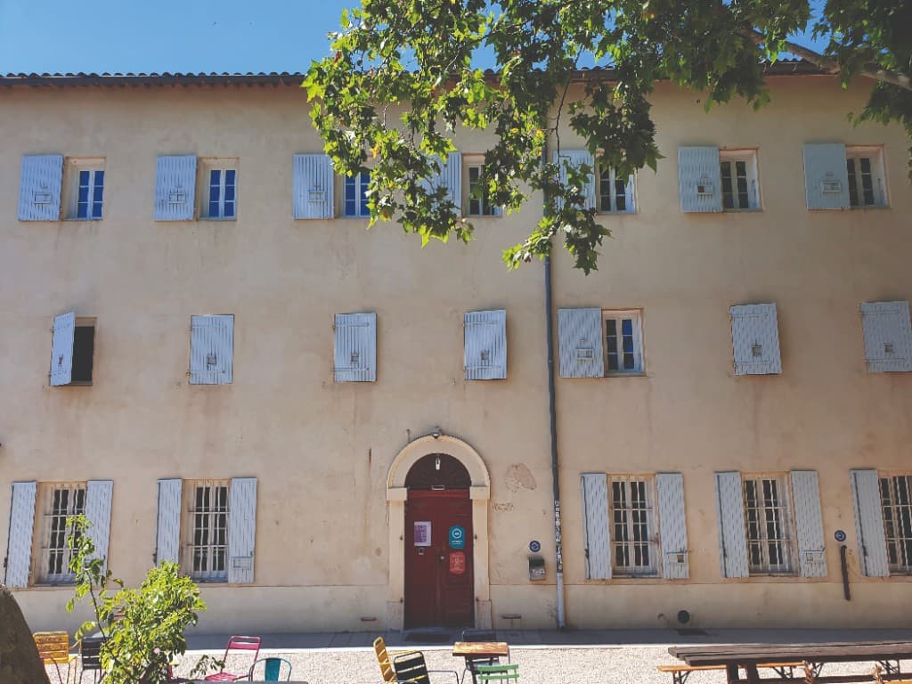 Façade ensoleillée du couvent Levat avec volets bleus et terrasse meublée de chaises colorées.